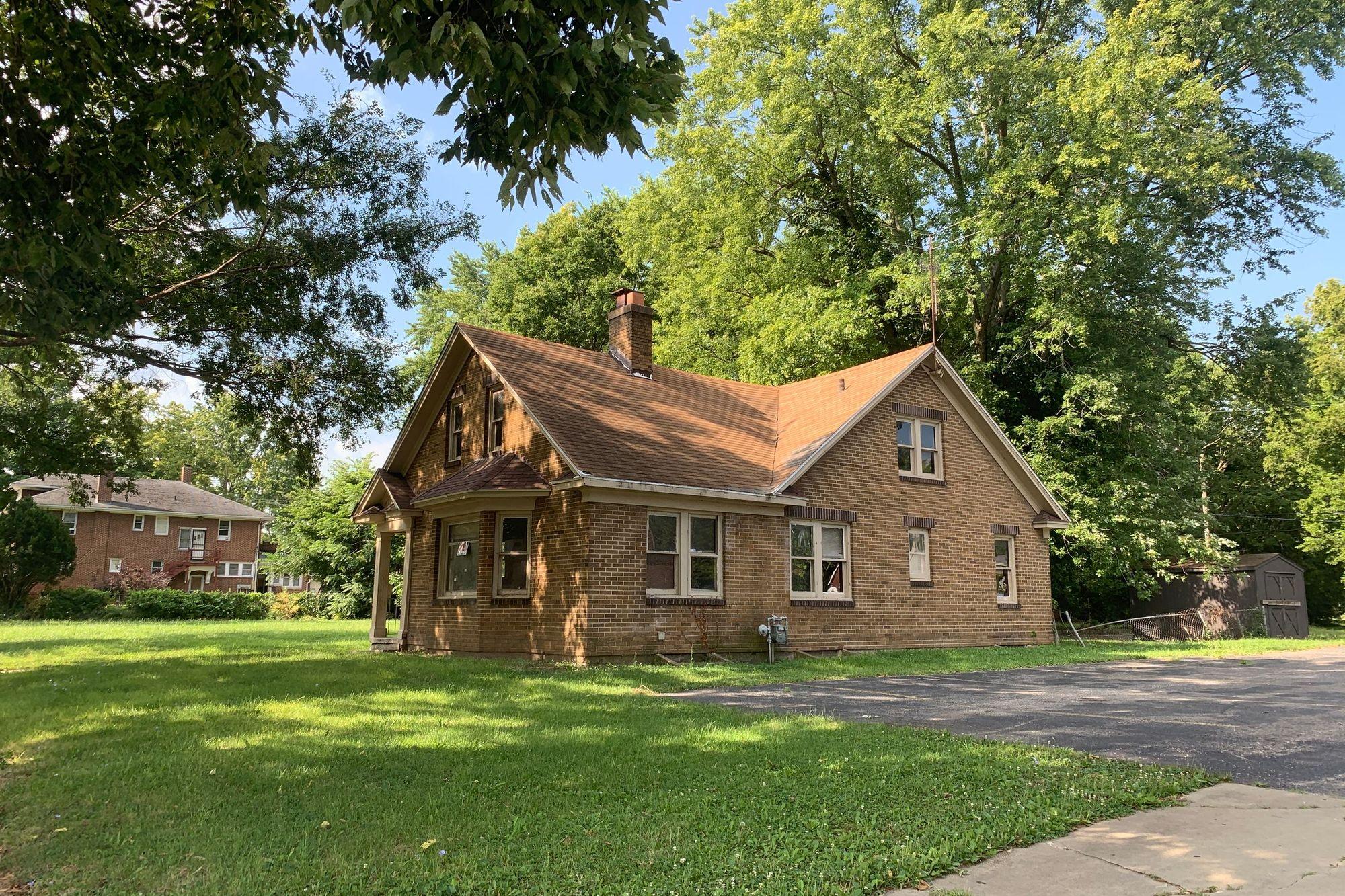 Older tan brick house on a large lawn under tall trees on a sunny summer day