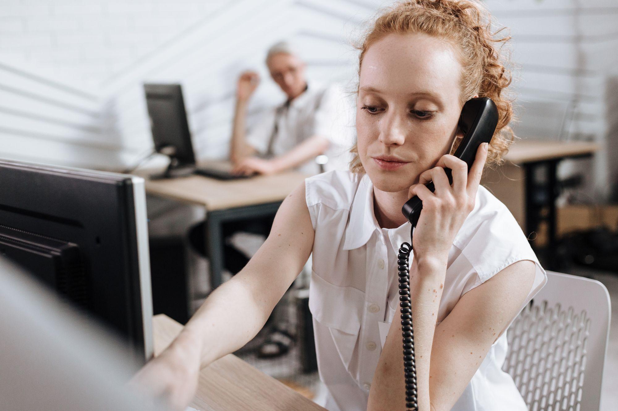 Woman on a corded office phone at her desk while a colleague works in the background