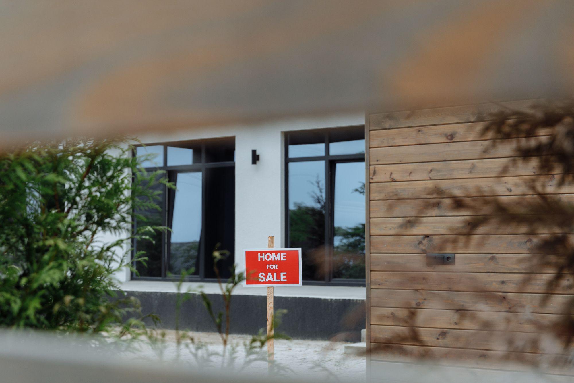 Modern house with a red Home for Sale sign in the front yard, framed by plants