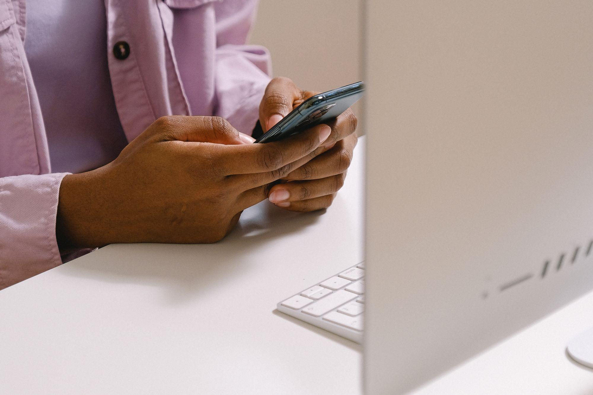 Hands typing a message on a smartphone at a white desk beside a computer keyboard
