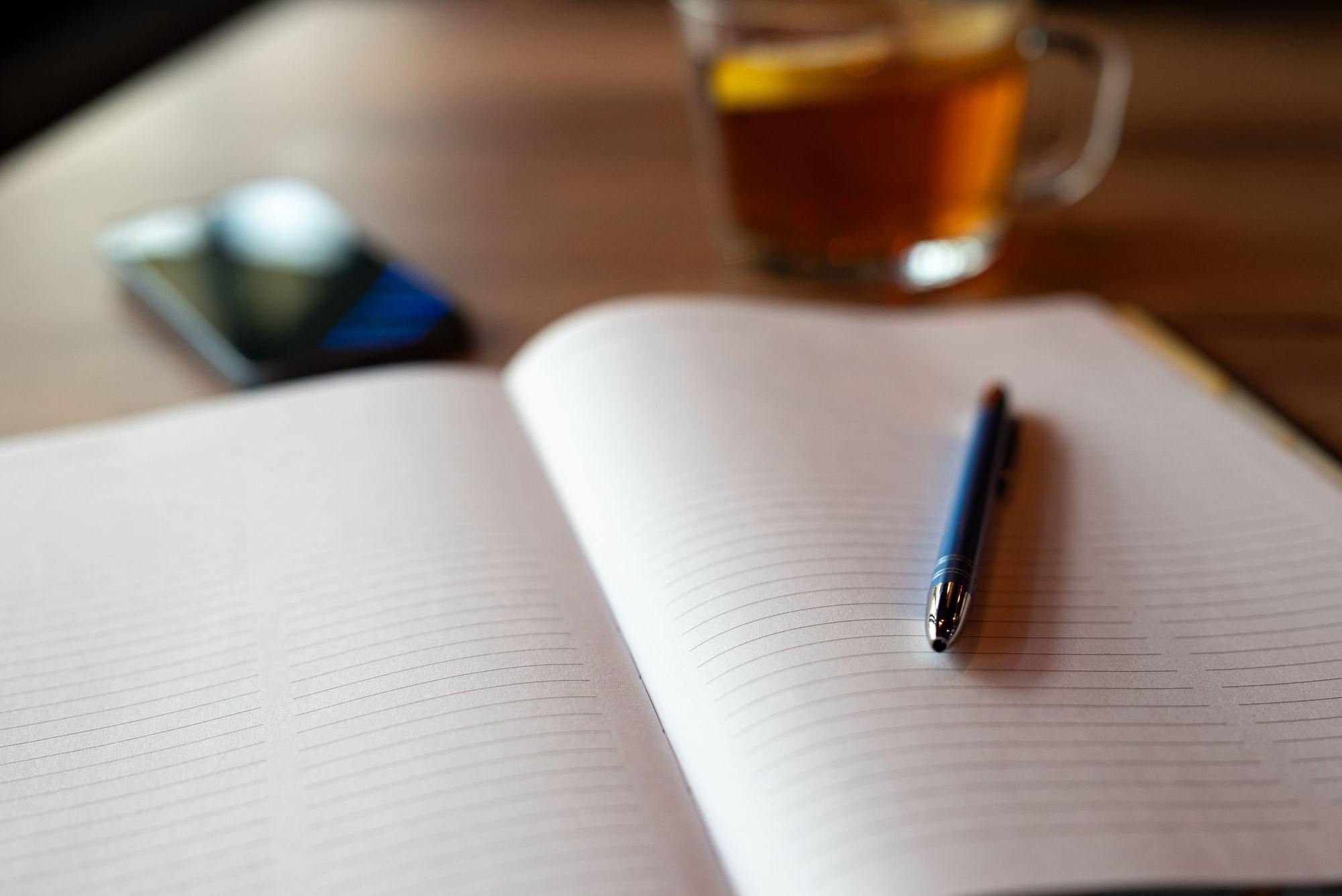 Open blank lined notebook with a blue pen, a phone and a glass of tea on a wooden table