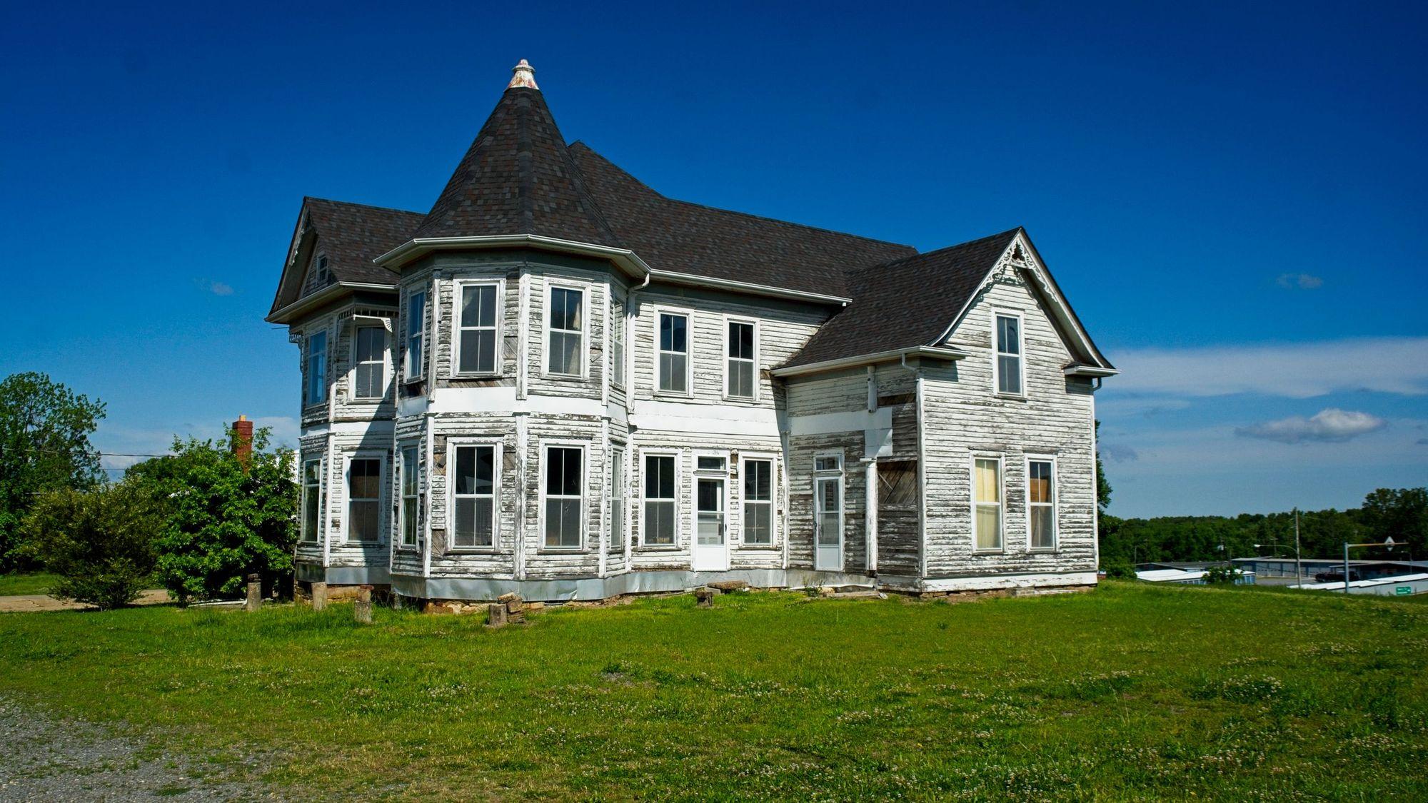 Weathered two-story Victorian house with peeling white siding on a grassy lot under a blue sky