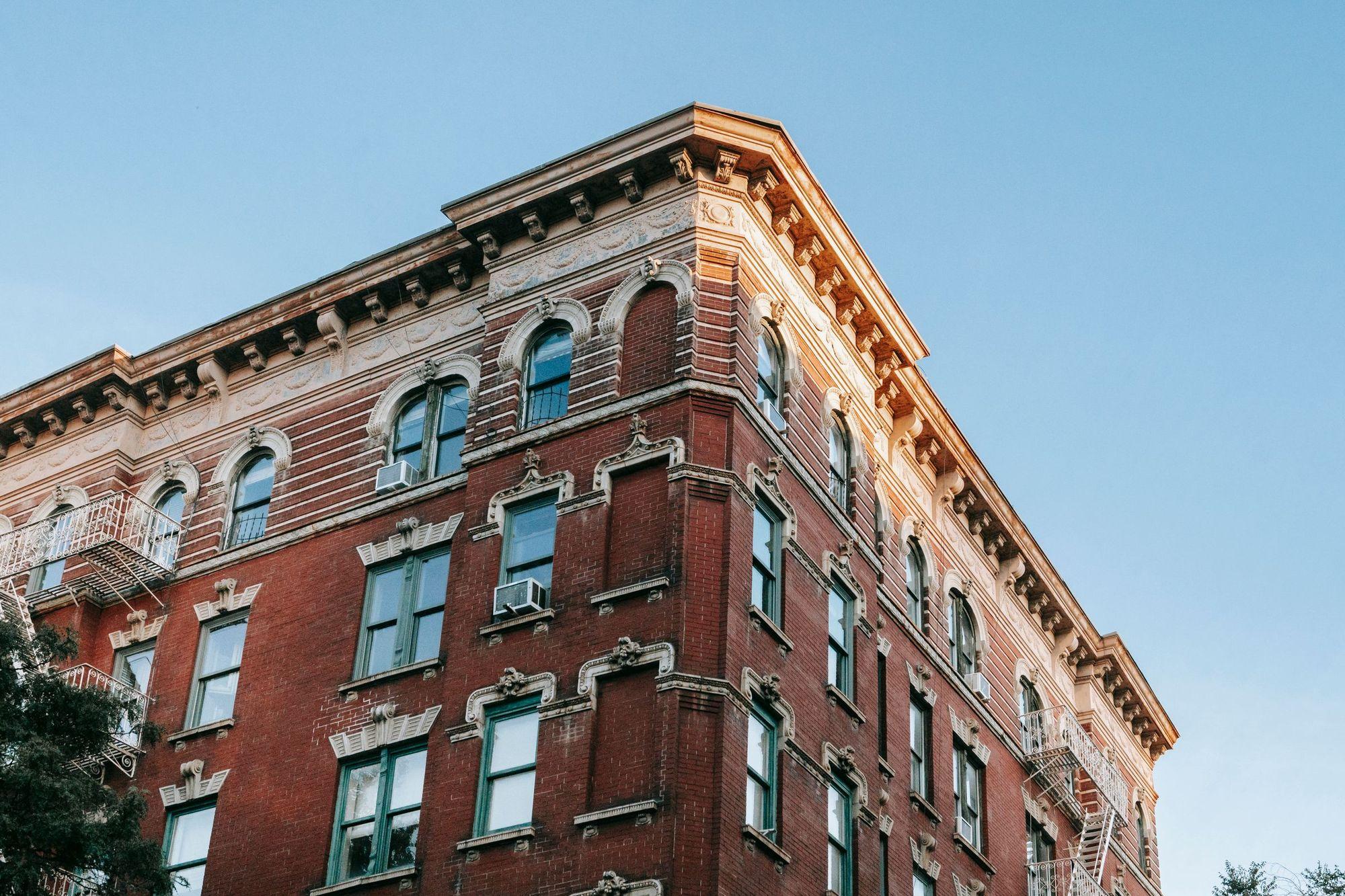 Corner of an old red brick walk-up apartment building with fire escapes under a clear blue sky
