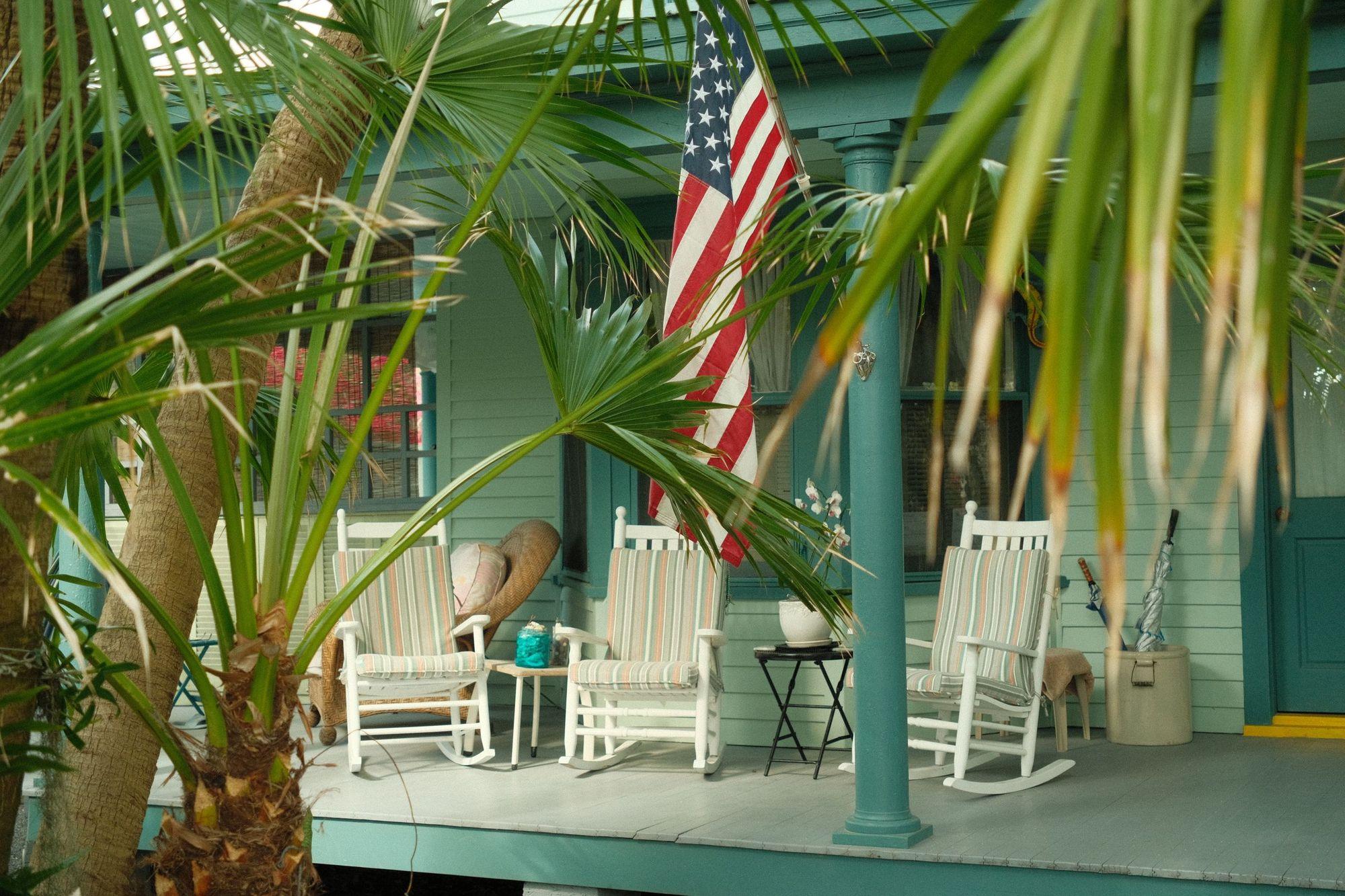 Front porch of an older wooden house with white rocking chairs, a potted orchid and an American flag