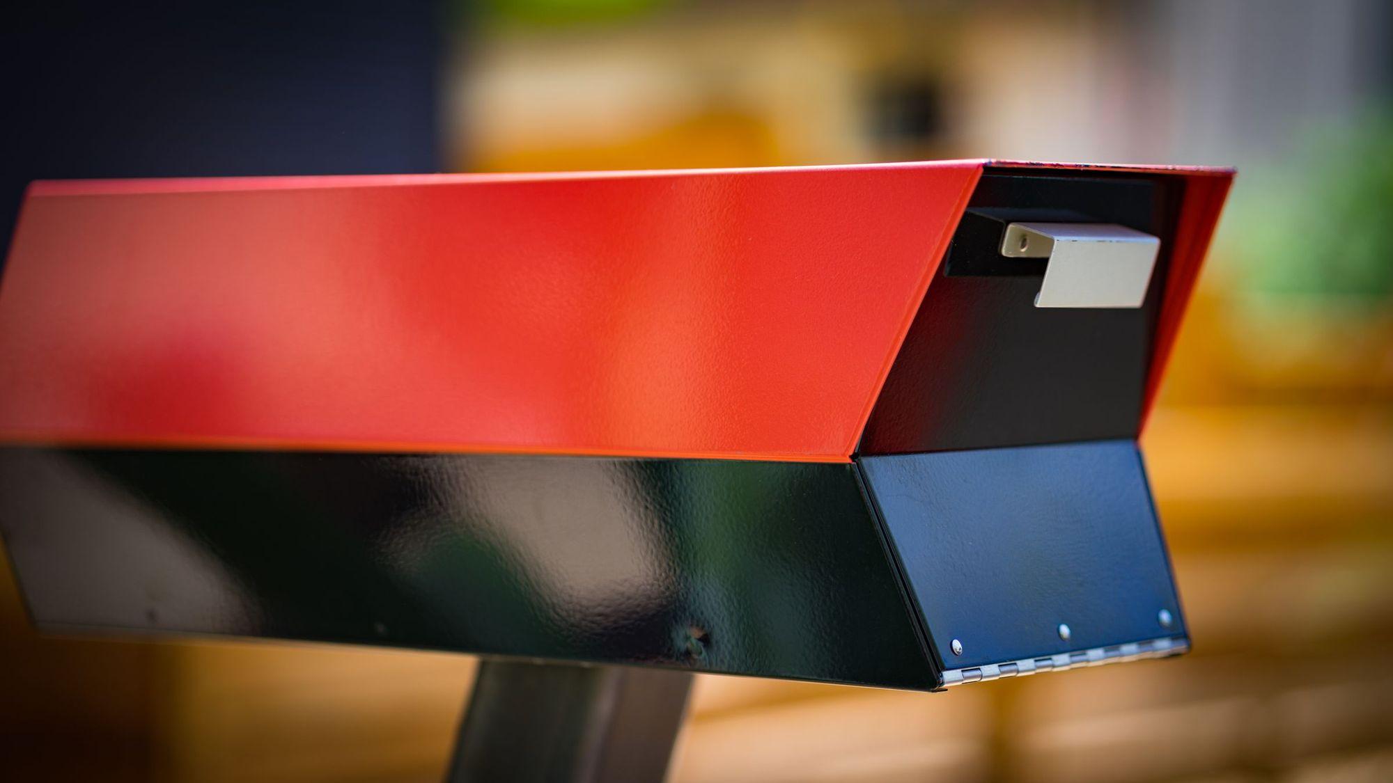 Close-up of a red and black metal mailbox on a post, a house blurred behind it