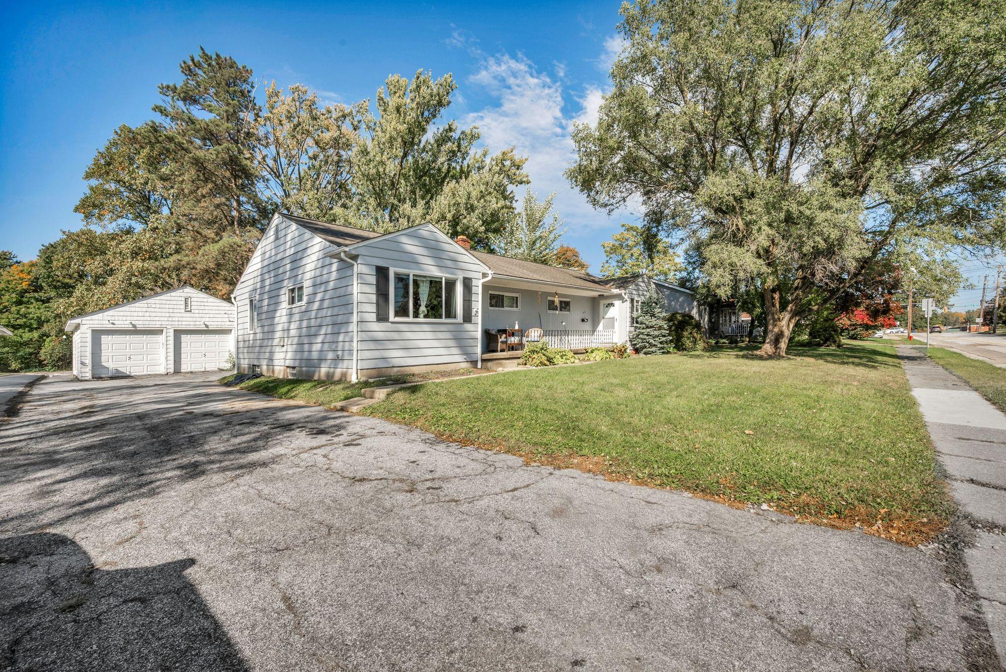 Dated single-story ranch house with detached garage and cracked driveway on a sunny suburban street