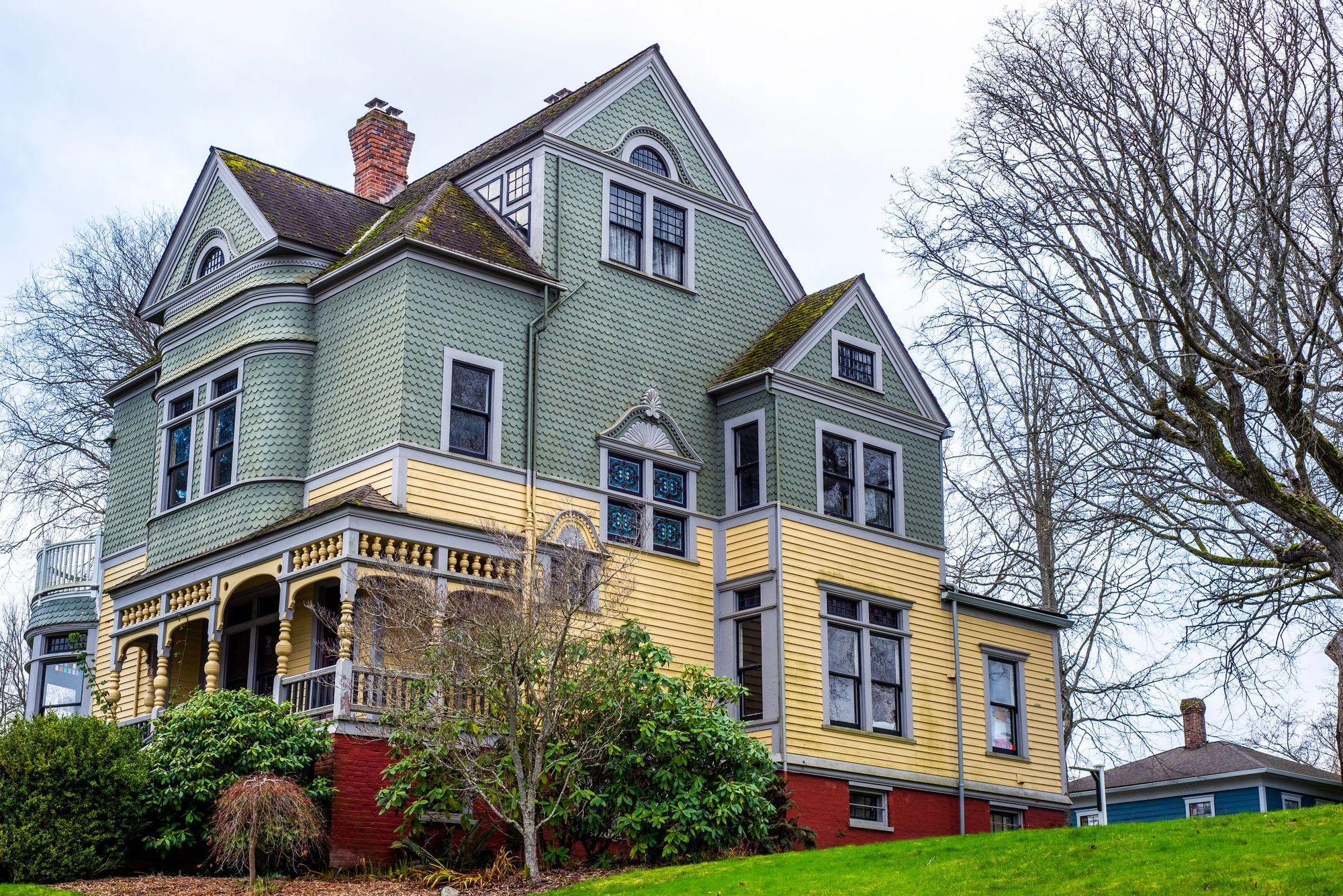 Older two-story Victorian house with a covered front porch, shrubs and a large bare tree