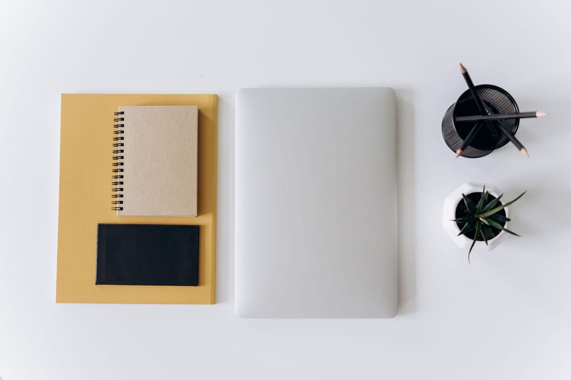 Closed silver laptop beside notebooks, a pencil cup and a small plant on a white desk