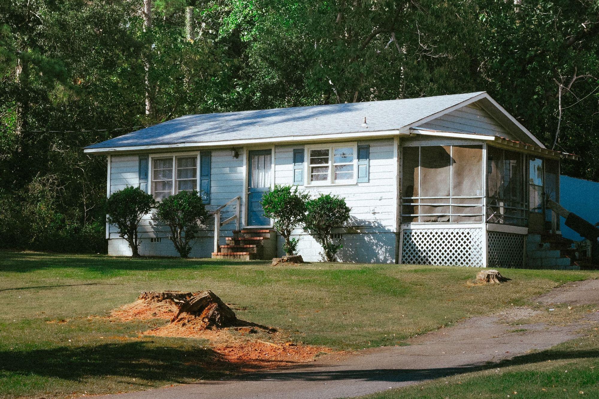 A worn single-story ranch house with a torn screen porch and tree stumps in the yard